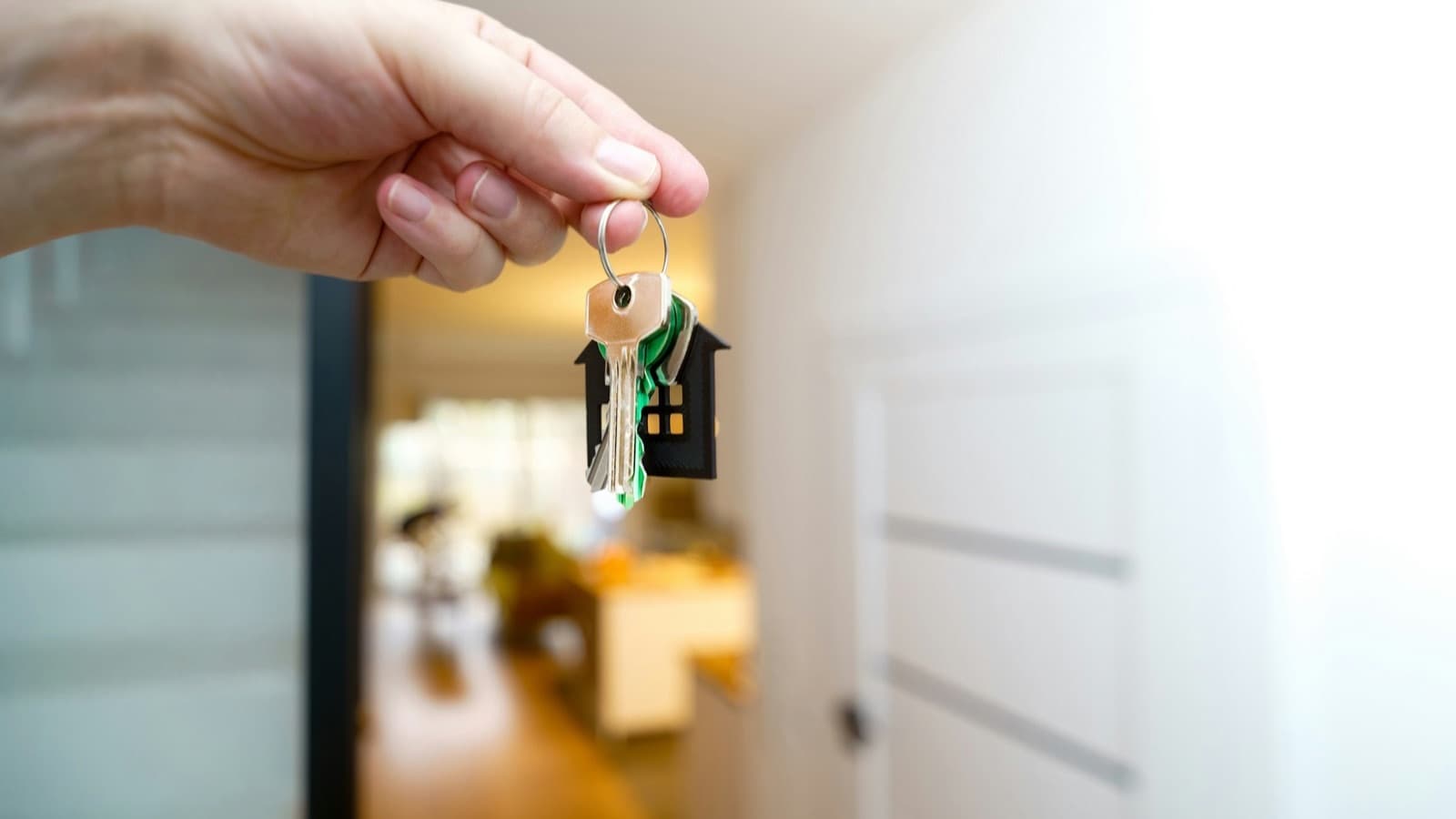 A hand holding a set of house keys on a ring with a small black house-shaped keychain, with a bright hallway softly blurred in the background